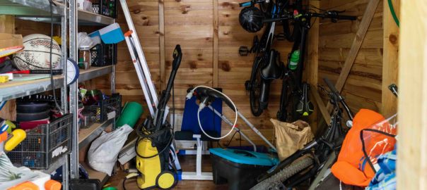Suburban home wooden storage utility unit shed with miscellaneous stuff on shelves, bikes, exercise machine, ladder, garden tools and equipment. Messy and chaos at house yard barn. Organization order
